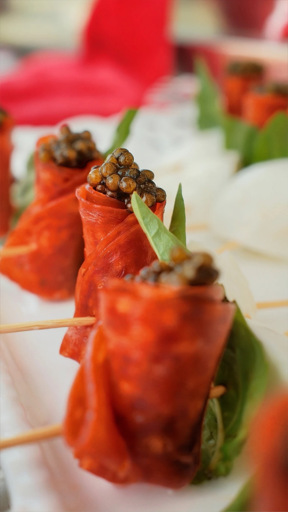 Pepperoni rolls on picks, each topped with golden caviar and a basil leaf, lined up on a white platter