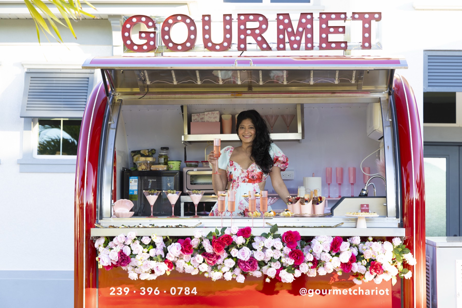 Nisha Singh at the Chariot window with a glass of champagne, beneath the GOURMET marquee and above the rose ledge